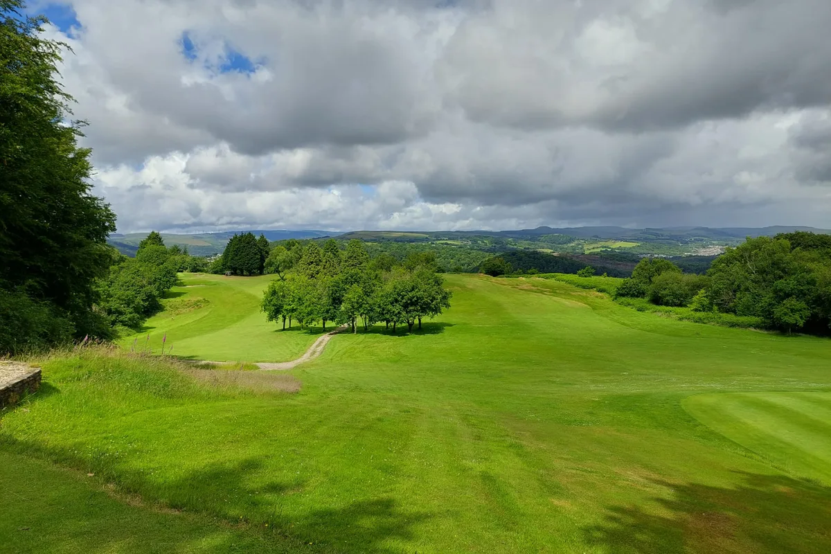 Looking down the fairway from the 5th tee, trees either side, the valley and hills beyond under a wide sky