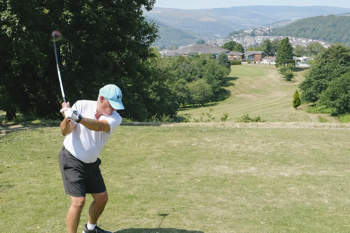 A golfer driving from a tee on the Common, with Pontypridd and the valley laid out below