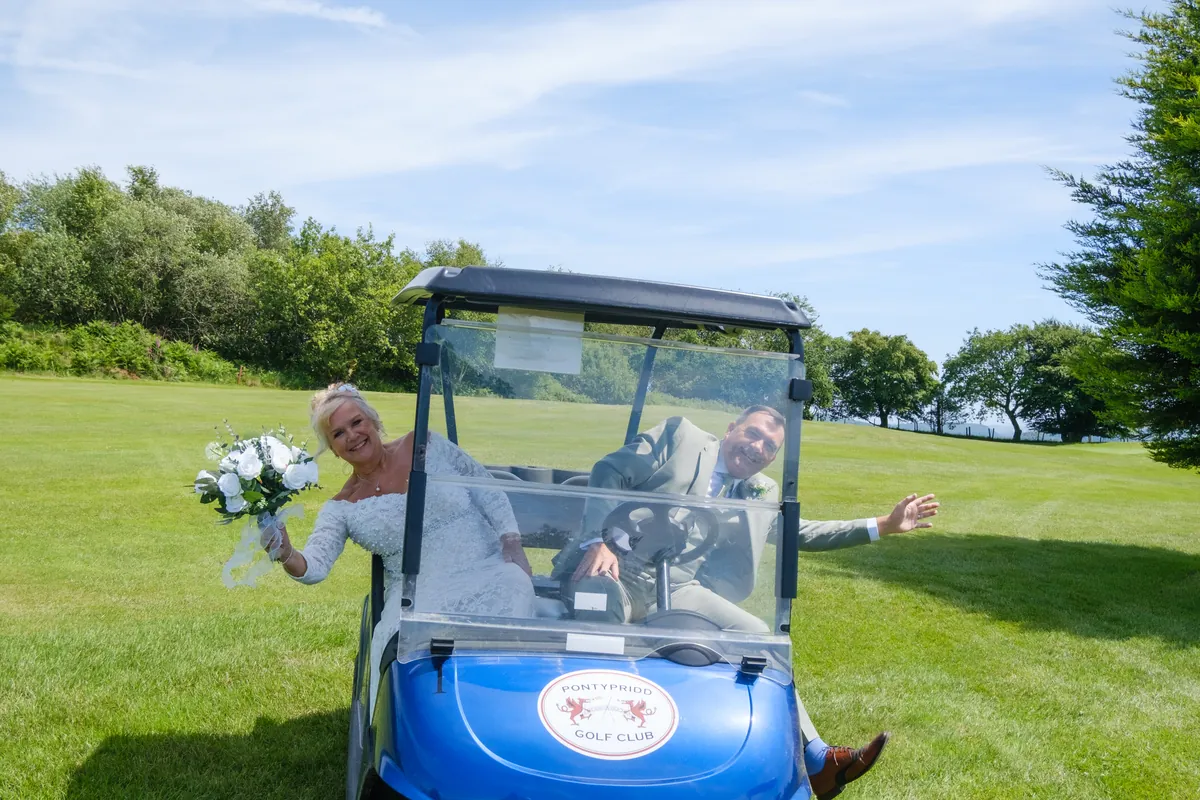 A bride riding a golf buggy across the course with her bouquet held out, arms wide