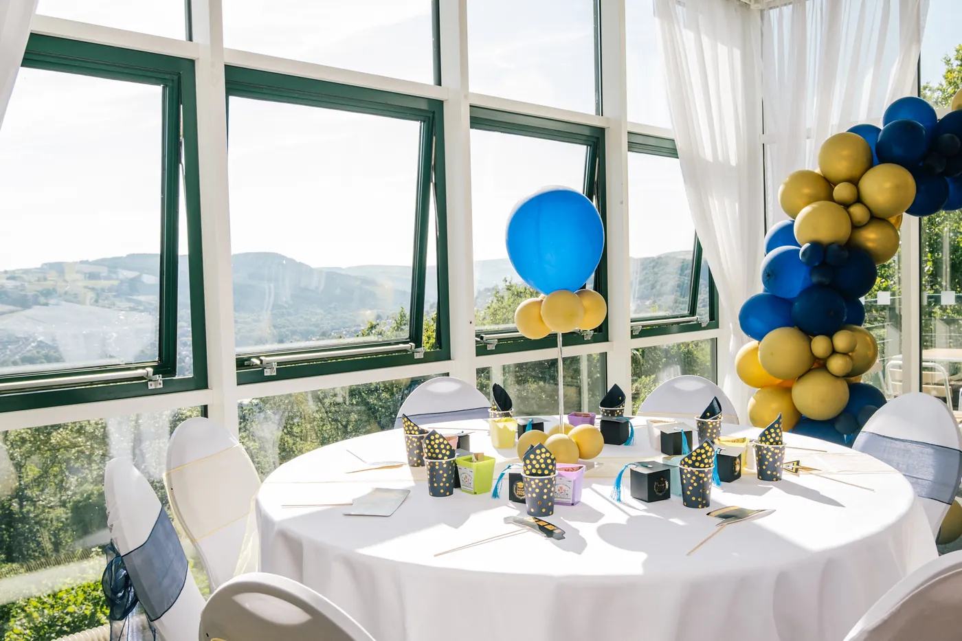 A round table dressed with a white cloth and a balloon arch, with the valley visible through the function room's floor-to-ceiling windows