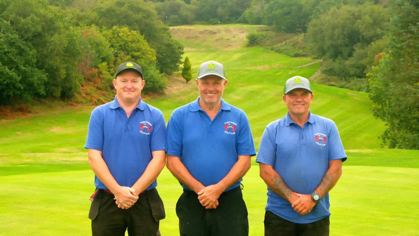 The three greenkeepers in club polo shirts and caps, standing on a green with a striped fairway climbing between the trees behind them