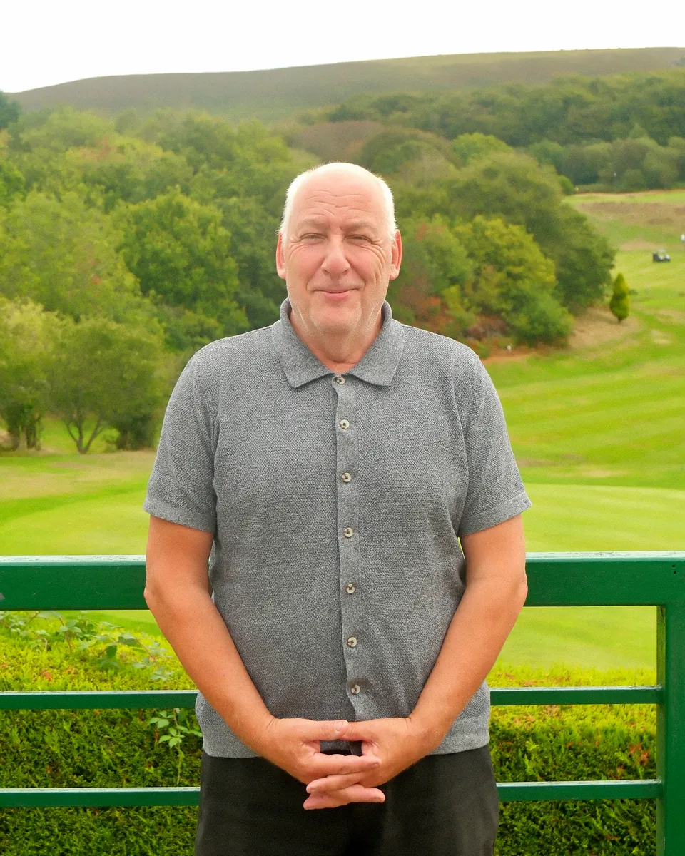 Peter Mattravers, the club secretary, standing at the green railing above the course, with fairways and wooded hills behind him