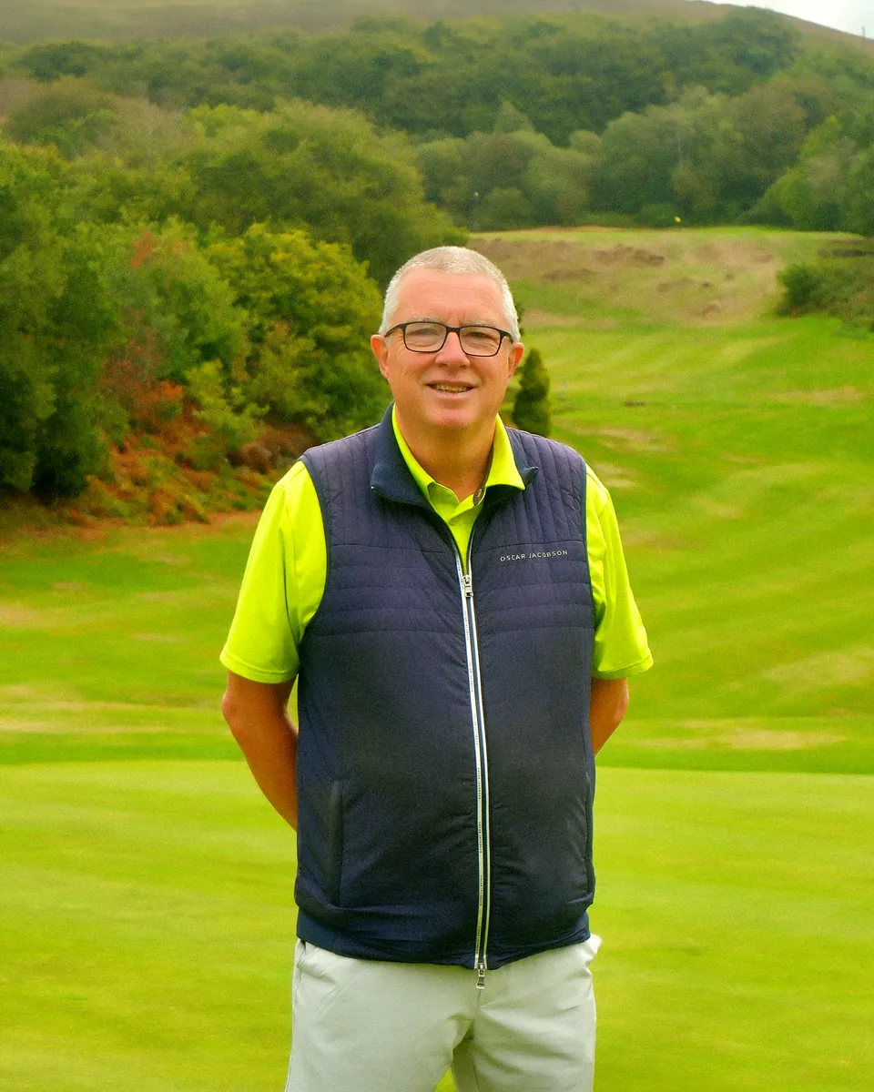 Tim Kelland, the club's new member ambassador, smiling on the course in a navy gilet, a striped fairway rising between the trees behind him
