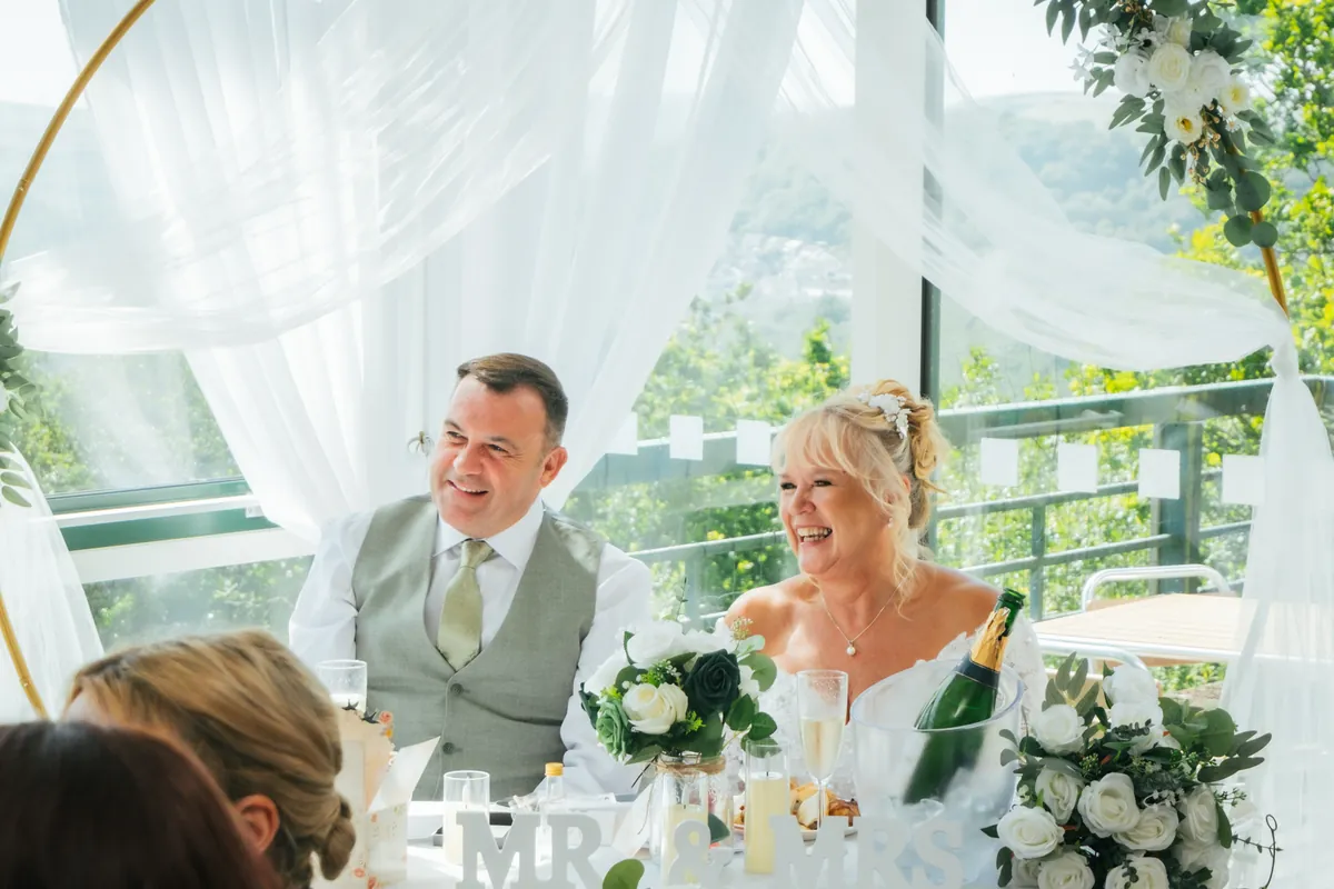 A newly married couple at the top table beneath a floral hoop, the windows and the view behind them